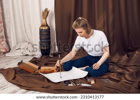 Cute relaxed blonde girl, painter or artist lying on a brown cloth in the comfy workshop, eating pizza and painting under the sun rays falling on her.
