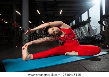 athletic woman in red sportswear sitting on yoga matte in black gym and warming up, girl doing yoga and stretching, attractive woman in fitness club doing flexibility exercise Royalty-Free Stock Photo #2310415115