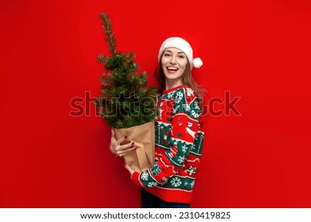 young cute girl in christmas sweater and santa hat holds artificial tree in shopping bag on red background, woman in christmas clothes bought tree and rejoice