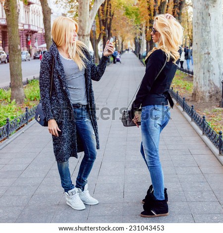 Two blonde sisters making photo shoot on smartphone in city avenue park in fall autumn sunny day, wearing trendy stylish casual clothes sneakers and sunglasses.