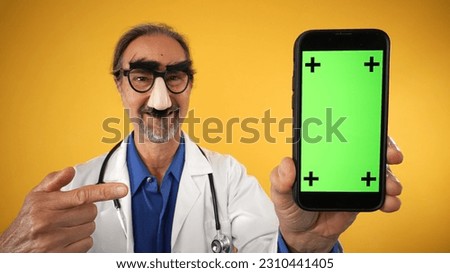 Mature elderly doctor man wear lab coat funny mustache glasses mask point finger on mobile cell phone with blank screen workspace area tracking points on solid yellow background studio portrait.
