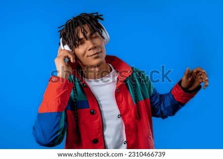 Positive young african american man listening music, enjoying dance with headphones on blue studio background. Radio, wireless modern sound technology, online player. High quality