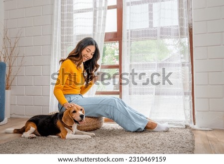 Young Asian girl sit on seat cushion also hold cup of tea and enjoy to play with beagle dog in front of glass door of the house with day light. Royalty-Free Stock Photo #2310469159