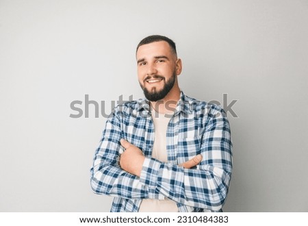 Confident young handsome man in shirt keeping arms crossed and smiling while standing against gray background