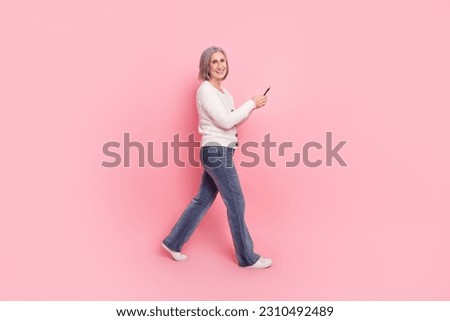 Full size photo of attractive senior woman walk hold gadget dressed stylish white clothes isolated on pink color background