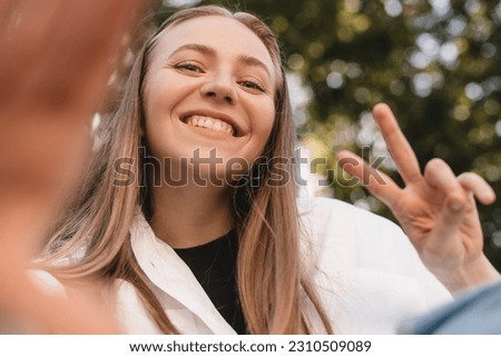 Pensive blonde woman make selfie posing on park background. Outdoor shot of happy business lady show peace sign. Business freedom style. Girl raises her hand to camera, show toothy smile, v gesture.