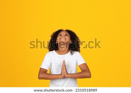 Smiling adolescent curly girl in white t-shirt looking up, praying gesture, isolated on yellow studio background. Dream come true, hope, study and education in school, ad, offer Royalty-Free Stock Photo #2310518989