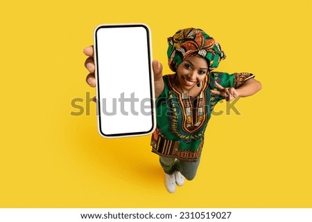 Smartphone with white blank screen in pretty black woman hand, happy young lady wearing national african costume showing great online deal and peace gesture, yellow studio background, mockup, top view Royalty-Free Stock Photo #2310519027