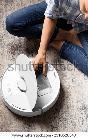 The girl takes the dust container out of the vacuum cleaner robot to clean it. The concept of a smart home, housework, cleaning, keeping the apartment clean, smart technologies. Royalty-Free Stock Photo #2310520343