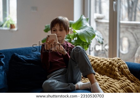 Relaxed little boy using mobile phone, looking at screen. Interested cute schoolboy sitting on couch at home with smartphone. Kid playing cellphone game, watch cartoons online. Child device addiction