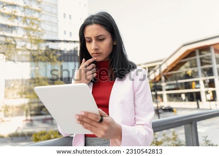 Serious attractive Indian woman holding digital tablet, check email, working online standing outdoors. Pensive asian student studying, learning language in university campus, education concept  Royalty-Free Stock Photo #2310542813