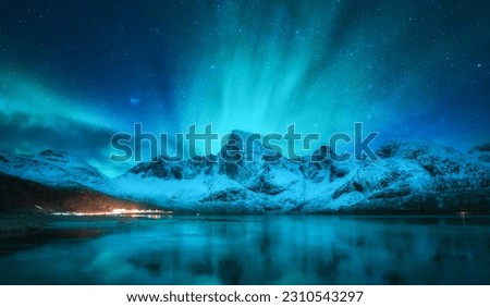 Northern lights over the snowy mountains, frozen sea coast, reflection in water at winter night in Lofoten, Norway. Aurora borealis and snow covered rocks. Nature. Polar lights, starry sky and fjord