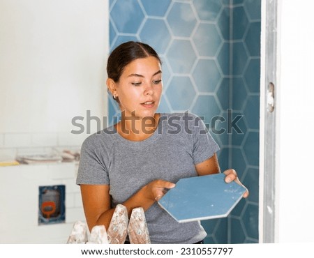 Young woman attentivelly examines the ceramic tile in her hands during renovation work in the bathroom Royalty-Free Stock Photo #2310557797