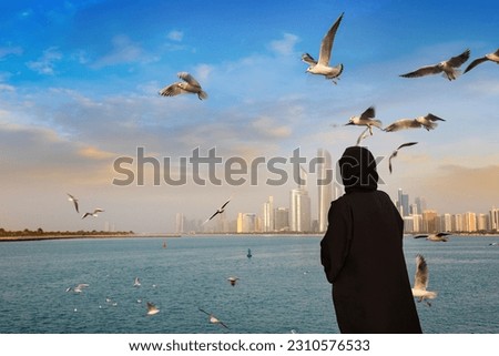 A woman wearing arab clothes is feeding seagulls in Abu Dhabi in a summer day, United Arab Emirates Royalty-Free Stock Photo #2310576533