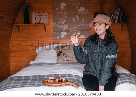 young caucasian woman with closed eyes woolen sweater and brown hat happy smiling at camera sitting on cozy log cabin bed before eating snack of small cereal toast and cheese, te wepu pods akaroa, new