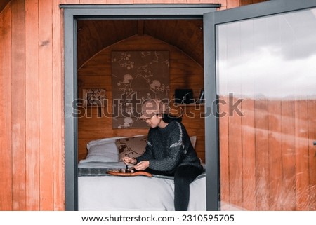 caucasian girl wearing brown hat and wool sweater sitting on bed spreading cheese on small cereal toast on small brown wooden board on bed inside small log cabin, te wepu pods akaroa, new zealand