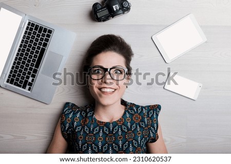 Overhead view of a young woman in glasses with emphatically enthusiastic expression, amidst tools for photography, graphic design, text publication, and online and traditional marketing campaigns. She