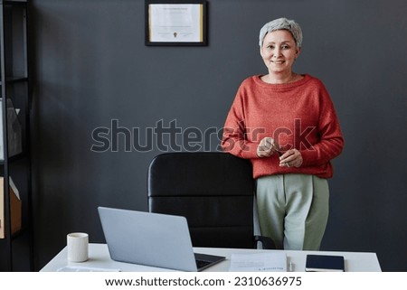 Portrait of successful senior woman standing by workplace in office wearing contrasting red outfit and smiling at camera, copy space