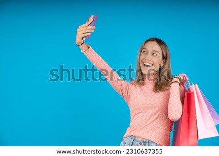 Smiling young woman taking a selfie and holding her gift bags, on a light blue background.