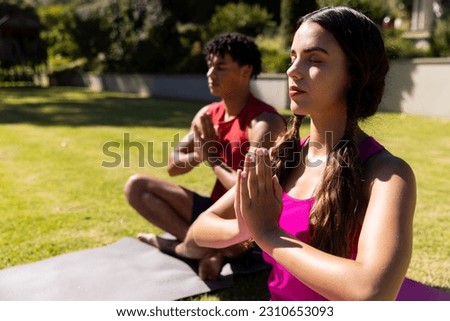 Biracial young woman with boyfriend practicing meditation in prayer pose while sitting in yard. Copy space, unaltered, love, togetherness, summer, zen, yoga, fitness and active lifestyle concept.