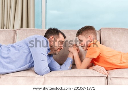 Handsome young father and his cute son competing in arm wrestling while lying on a sofa. Father and son quality time