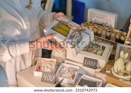 candle maker shares her creation with an eager buyer, surrounded by an array of fragrant candles and artisanal products at a vibrant fair. Sustainable Shopping: Supporting Local Artisans Royalty-Free Stock Photo #2310742973