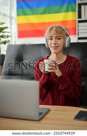 Portrait of a creative and confident young Asian gay man freelancer is working from home, holding a coffee mug and listening to music through headphones in the living room.