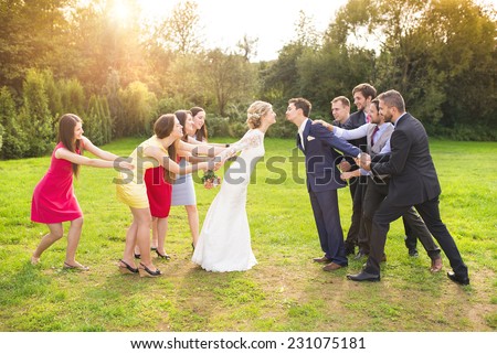 Funny portrait of newlywed couple kissing, bridesmaids and groomsmen pulling them away in green sunny park