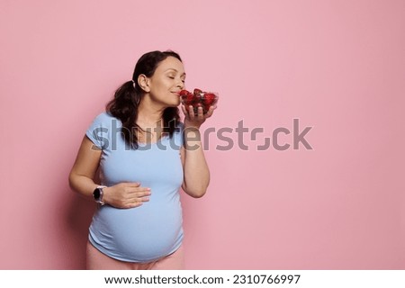 Attractive conscious pregnant woman puts her hand on her big belly in late pregnancy, enjoying the smell of delicious organic strawberries in a glass bowl, isolated on pink background. Copy ad space