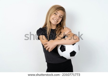 Charming pleased beautiful caucasian teen girl wearing sportswear holding a football ball over white wall embraces own body, pleasantly feels comfortable poses. Tenderness and self esteem concept Royalty-Free Stock Photo #2310771773