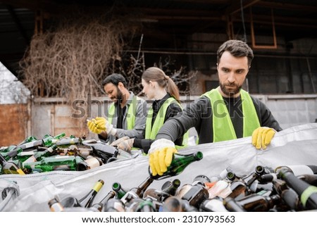 Bearded worker in protective gloves and vest putting glass bottle in sack near blurred interracial colleagues in outdoor waste disposal station, garbage sorting and recycling concept Royalty-Free Stock Photo #2310793563