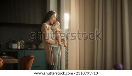 Beautiful Young Asian Woman Holding her Baby in her Arms While Standing Next to a Window at Home. Cute Little Toddler Resting in His Mother's Embrace as She Shows him the Busy Streets of Urban City. Royalty-Free Stock Photo #2310793913