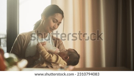 Portrait of Beautiful Asian Mother Feeding Milk to her Infant Using a Baby Bottle at Bright Home. Young Woman New to Motherhood Bonding with her Child and Enjoying an Affectionate Family Moment. Royalty-Free Stock Photo #2310794083