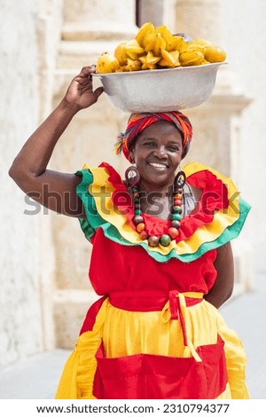 Cheerful fresh fruit street vendor aka Palenquera in the Old Town of Cartagena de Indias, Colombia. Happy, smiling Afro-Colombian woman in traditional clothing, Colombian culture and lifestyle.  Royalty-Free Stock Photo #2310794377