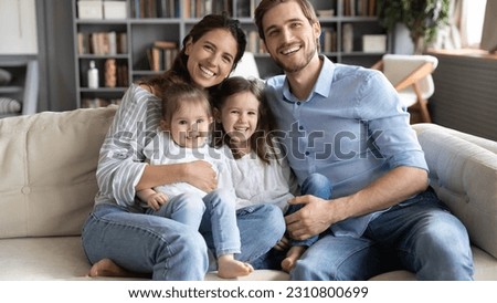 Portrait of happy young Caucasian family with two little girls children sit on couch in living room relaxing. Smiling parents with small daughters rest on sofa at home enjoy leisure weekend together.
