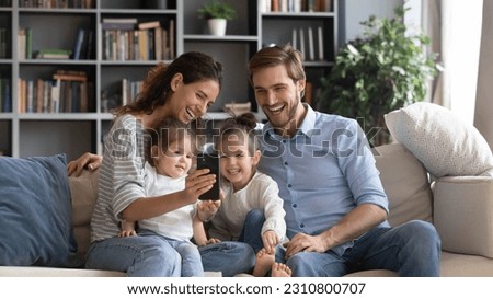 Overjoyed young Caucasian family with two small daughters relax on couch at home talk on video call on cellphone. Happy parents with little girls children use modern smartphone gadget together.