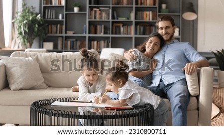 Little cute Caucasian girls children have fun draw paint in cozy home, happy young parents relax on couch together. Smiling family with two small daughters kids rest on leisure weekend in living room.