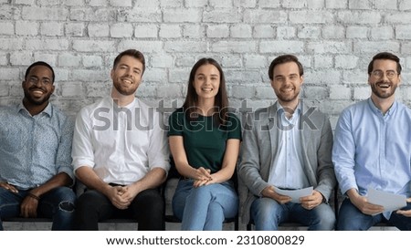 Confident diverse vacancy applicants sitting in queue, looking at camera, smiling. Happy job candidates waiting for interview in corridor, holding paper resume, ready for appointment. Group portrait