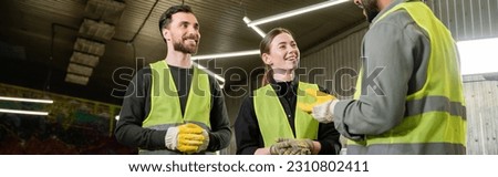 Cheerful workers in protective gloves and high visibility vests looking at indian colleague talking while standing together in blurred waste disposal station, recycling concept, banner