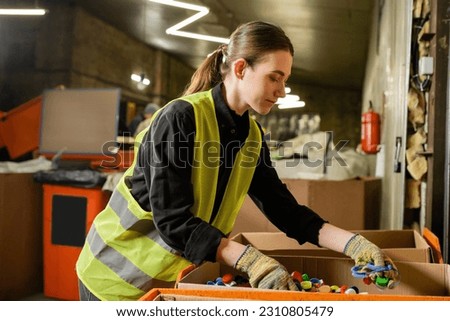Young female worker of garbage sorting center wearing protective clothing and gloves while working with plastic caps in carton boxes, garbage sorting and recycling concept Royalty-Free Stock Photo #2310805479