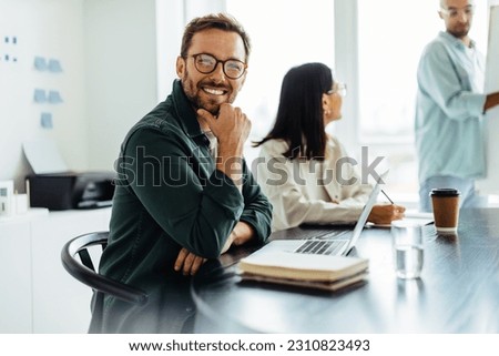 Business man smiling at the camera while sitting in a boardroom during a meeting. Happy business professional sitting in a presentation with a laptop.