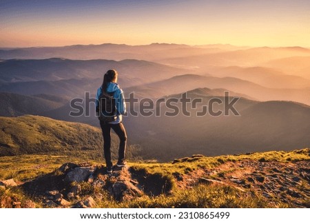 Girl with backpack on mountain peak with green grass looking in beautiful mountain valley in fog at sunset in autumn. Landscape with sporty young woman, foggy hills, orange sky in fall. Hiking. Nature Royalty-Free Stock Photo #2310865499