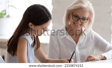Smiling young Caucasian female employee put signature on contract close deal with middle-aged employer in office. Happy diverse businesswoman sign document make agreement after successful meeting.
