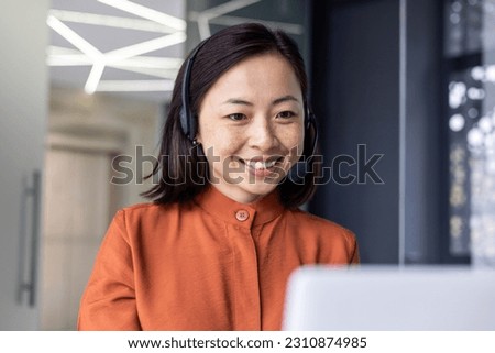 Close-up photo of young beautiful Asian woman talking on video call, looking at laptop web camera and smiling using headset for communication, successful businesswoman working in office. Royalty-Free Stock Photo #2310874985