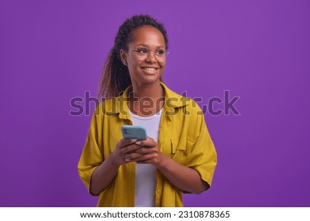 Young attractive happy African American woman with mobile phone looking away coming up with SMS to send to boyfriend or witty personal blog status stands posing in purple studio. Chatting, messenger