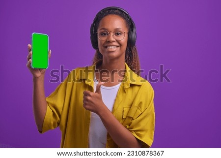 Young positive ethnic African American woman in headphones demonstrating green screen phone and showing thumb up recommending good mobile app or website stands on purple studio background.