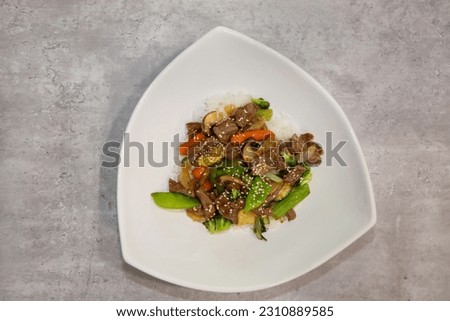 Beef stir-fry with vegetables served on top of steamed white rice in a white bowl. 
