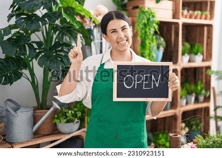 Young hispanic woman working at florist holding open sign smiling with an idea or question pointing finger with happy face, number one 