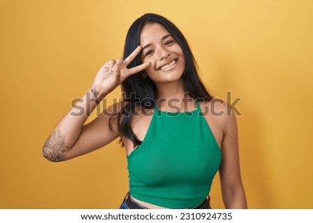 Brunette woman standing over yellow background doing peace symbol with fingers over face, smiling cheerful showing victory 