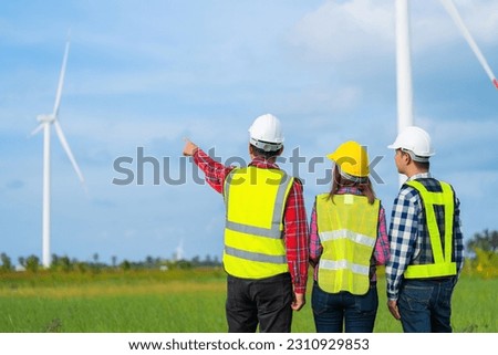 photo of the back of a team of 3 Asian male and female engineers pointing to a destination working together There is a wind turbine spinning as an energy industry business wearing a vest and helmet.